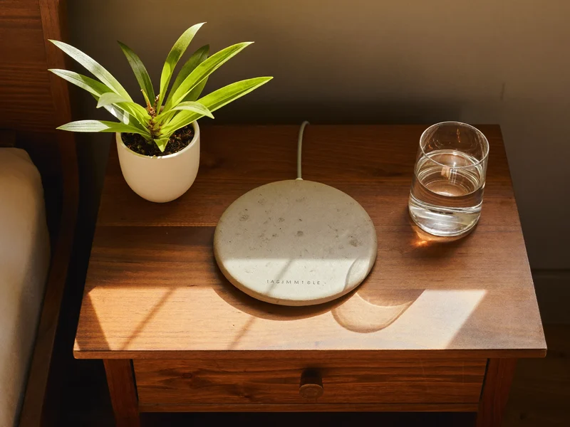 Încărcător wireless rotund, cu finisaj cald de piatră, pe o noptieră din lemn, lângă o plantă